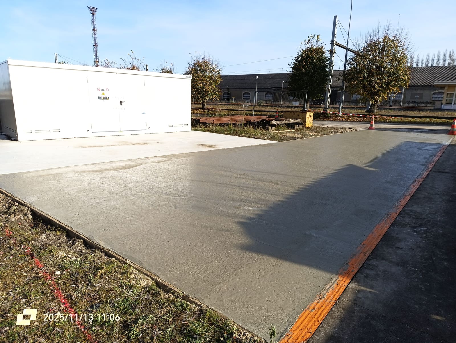 Dalle en béton armé fraîchement coulée et talochée devant un local technique, voies et caténaires à l’arrière-plan