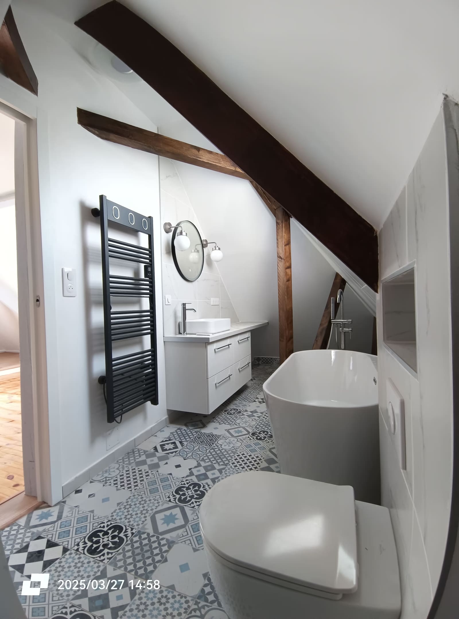 Salle de bain aménagée sous rampants : baignoire îlot, vasque suspendue, carreaux de ciment et poutre apparente